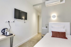 Una habitación de hotel moderna con un televisor de pantalla plana montado en la pared, una mesita de noche con una tetera y una taza, una cama con almohadas blancas y marrones, y una lámpara de pared redonda.