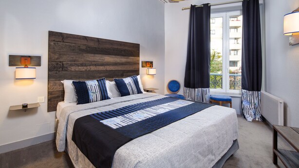 A modern bedroom with a large bed, wooden headboard, bedside lamps, a window with curtains, and a view of a building outside. - Hôtel Claret Bercy (Paris)