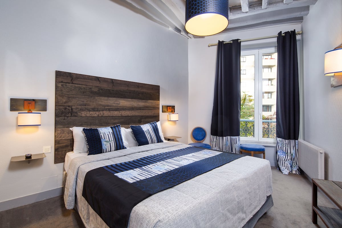 A bedroom with a large bed, a wooden headboard, bedside lamps, a window with curtains, and a view of a building outside.