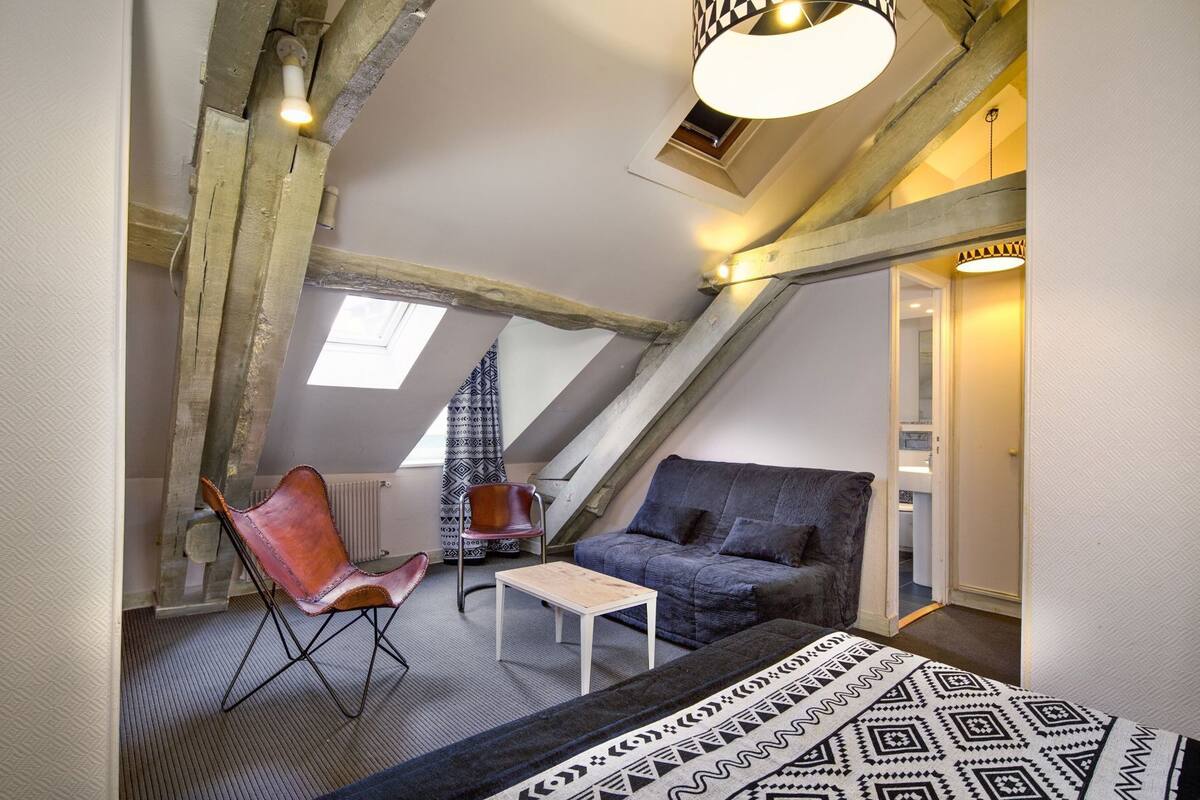 A loft bedroom with exposed wooden beams, a sloped ceiling, a window, a sofa, a chair, a small table, and a patterned rug.