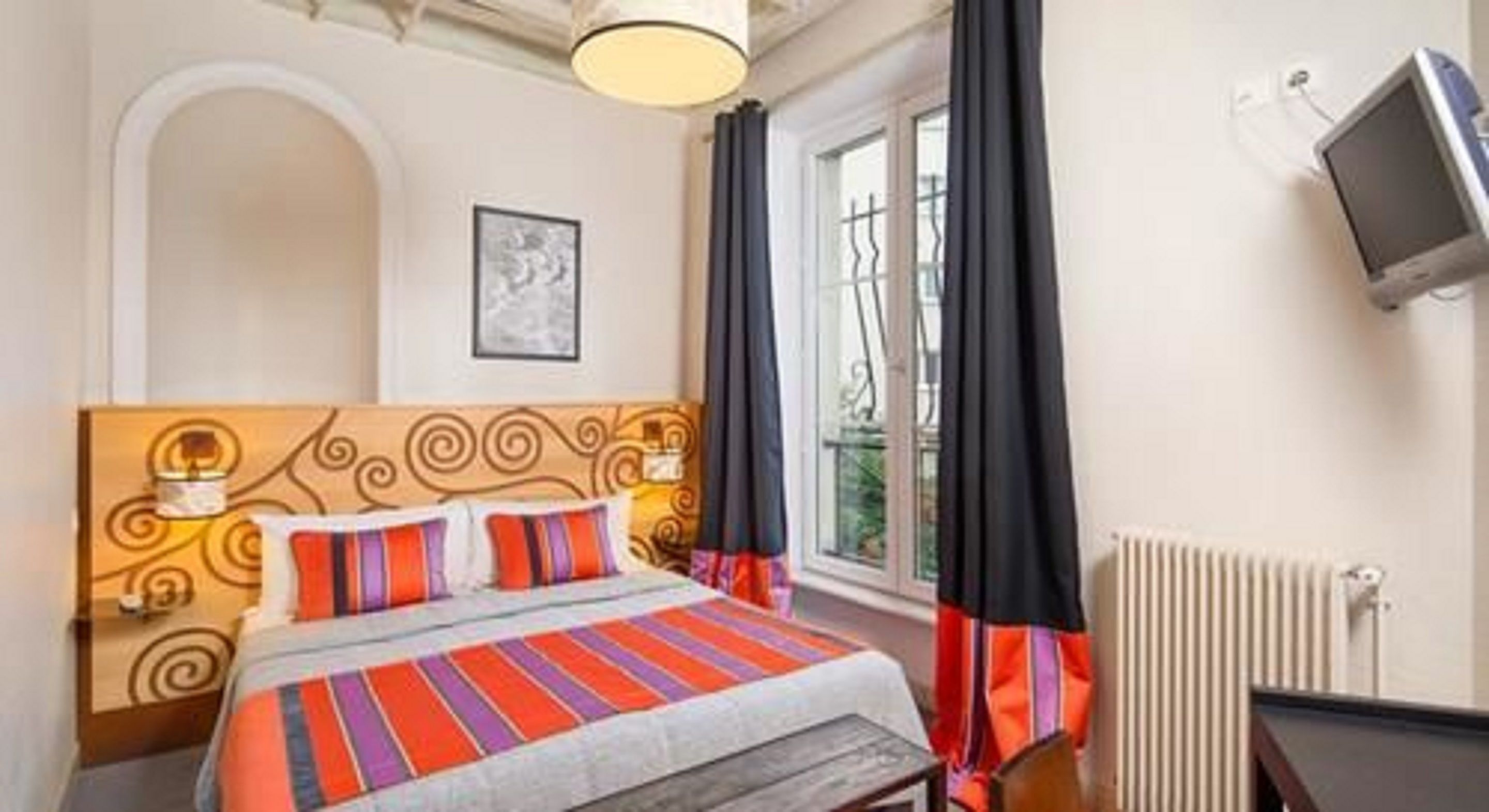 Une chambre d’hôtel comprenant un lit, un bureau, une chaise, une fenêtre avec des rideaux, un radiateur et un téléviseur fixé au mur.