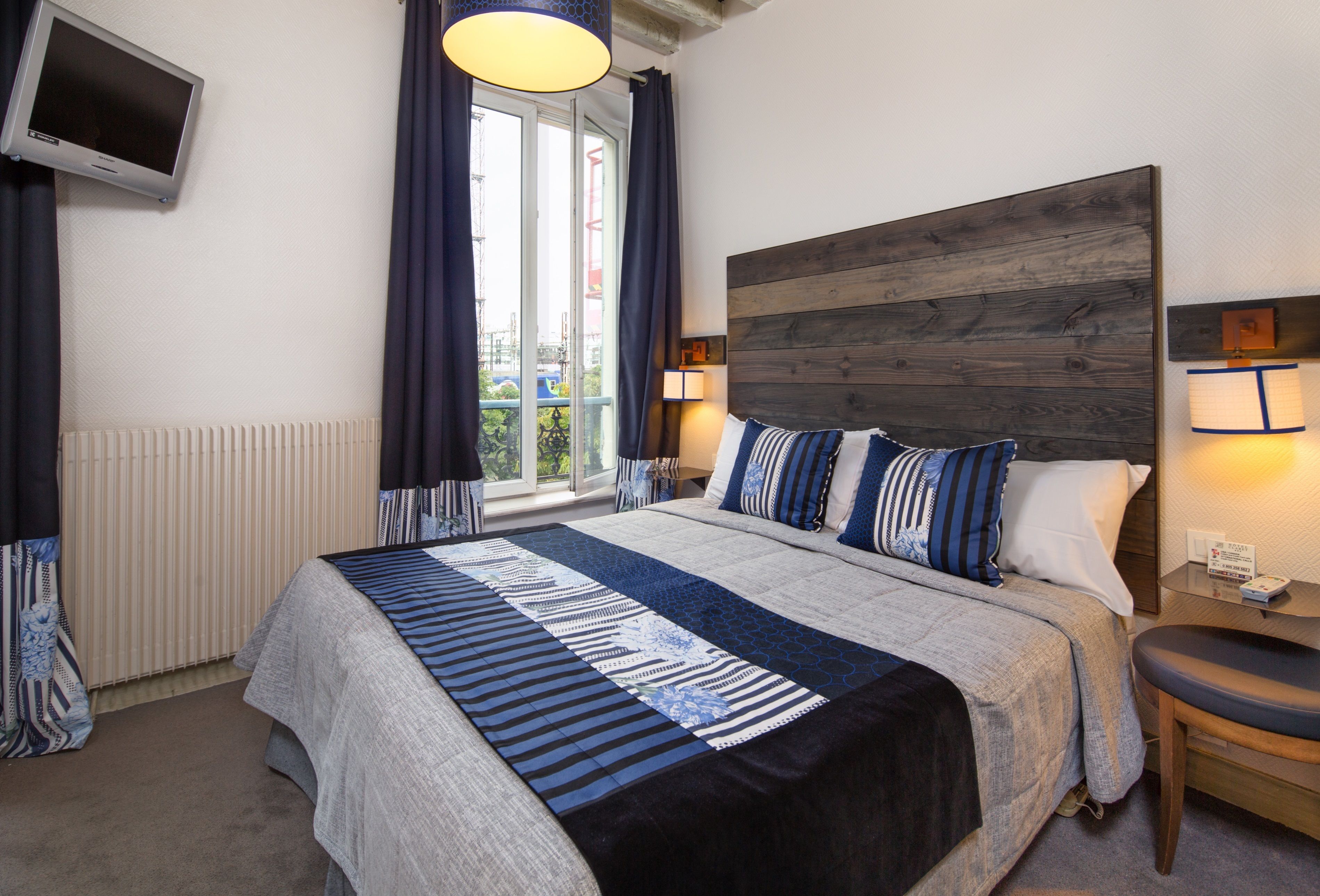 Habitación de hotel con una cama grande, cabecera de madera, dos lámparas de noche, televisor montado en la pared y una ventana con cortinas.