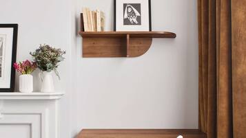Een hoekkamer met een witte schoorsteen, een houten bureau met een stoel, een plank met boeken en een lijst met een afbeelding, en een raam met bruine gordijnen.