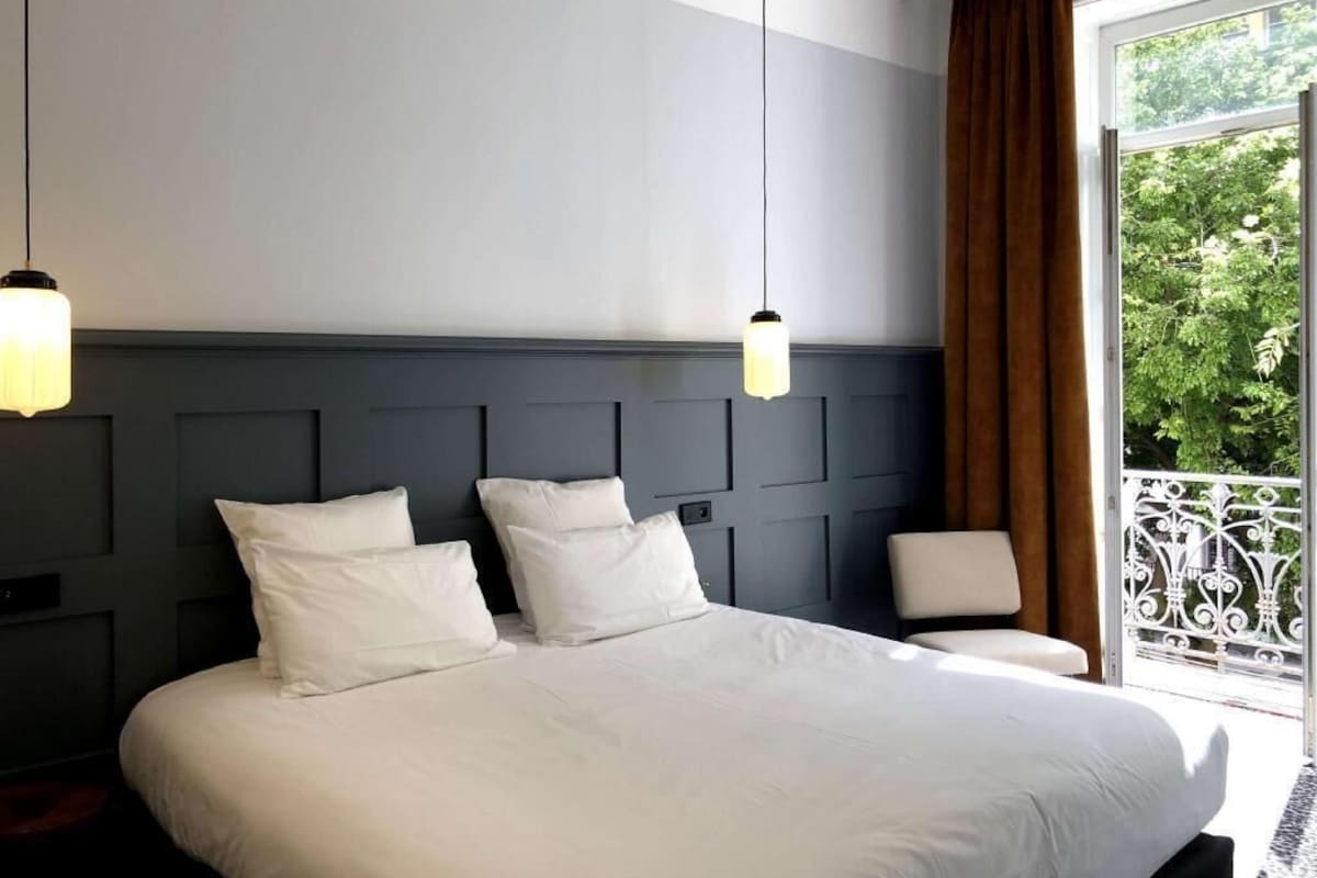A hotel room with a large bed, a headboard with a geometric design, two bedside lamps, and a view of greenery through a balcony.