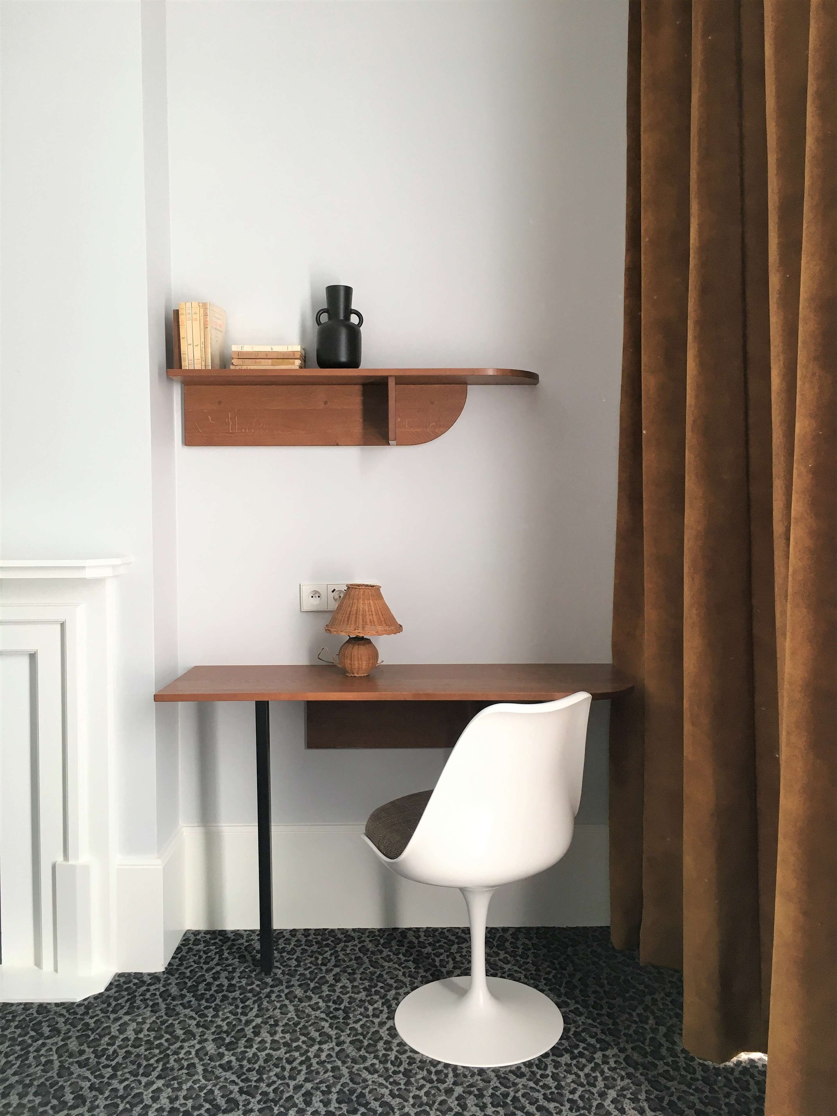 Escritorio en esquina con una silla blanca, una lámpara de fibras naturales y dos estantes de madera con libros y un jarrón negro.