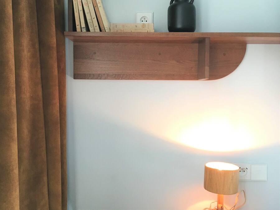 Estante de madeira com caneca preta e livros, luminária em uma mesa abaixo e uma tomada na parede.