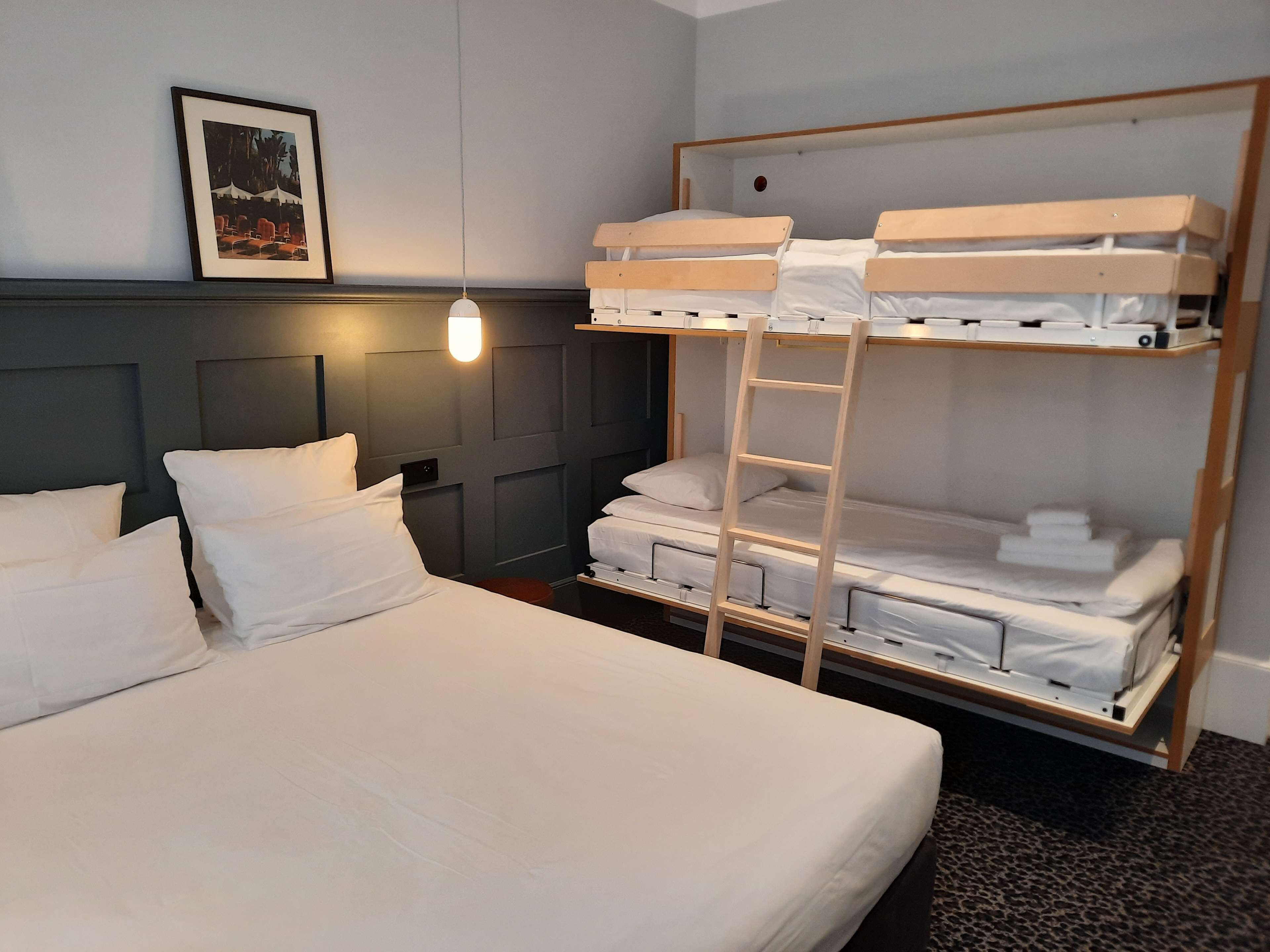 A hotel room with a bunk bed, a single bed, a framed picture on the wall, and a bedside lamp.