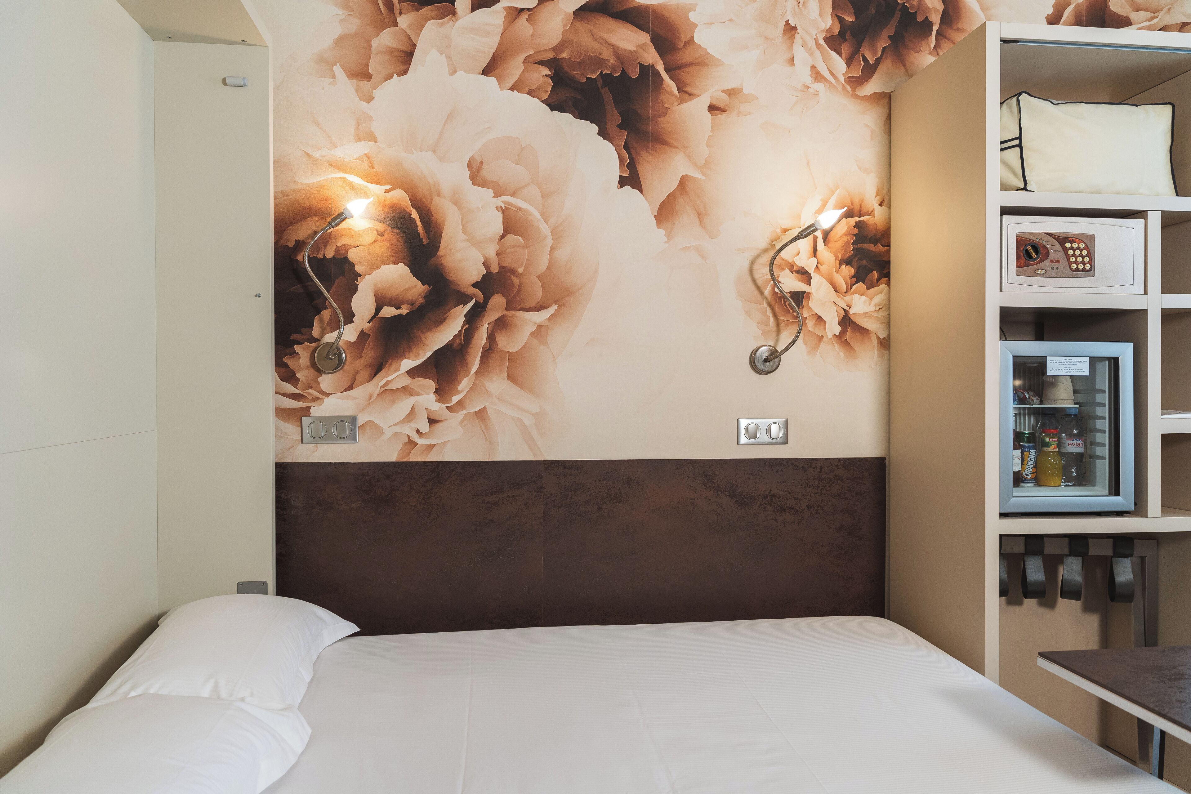 Une chambre d’hôtel avec un grand décor mural représentant des motifs floraux, un lit avec du linge de maison blanc, une table de chevet et un mini-bar.