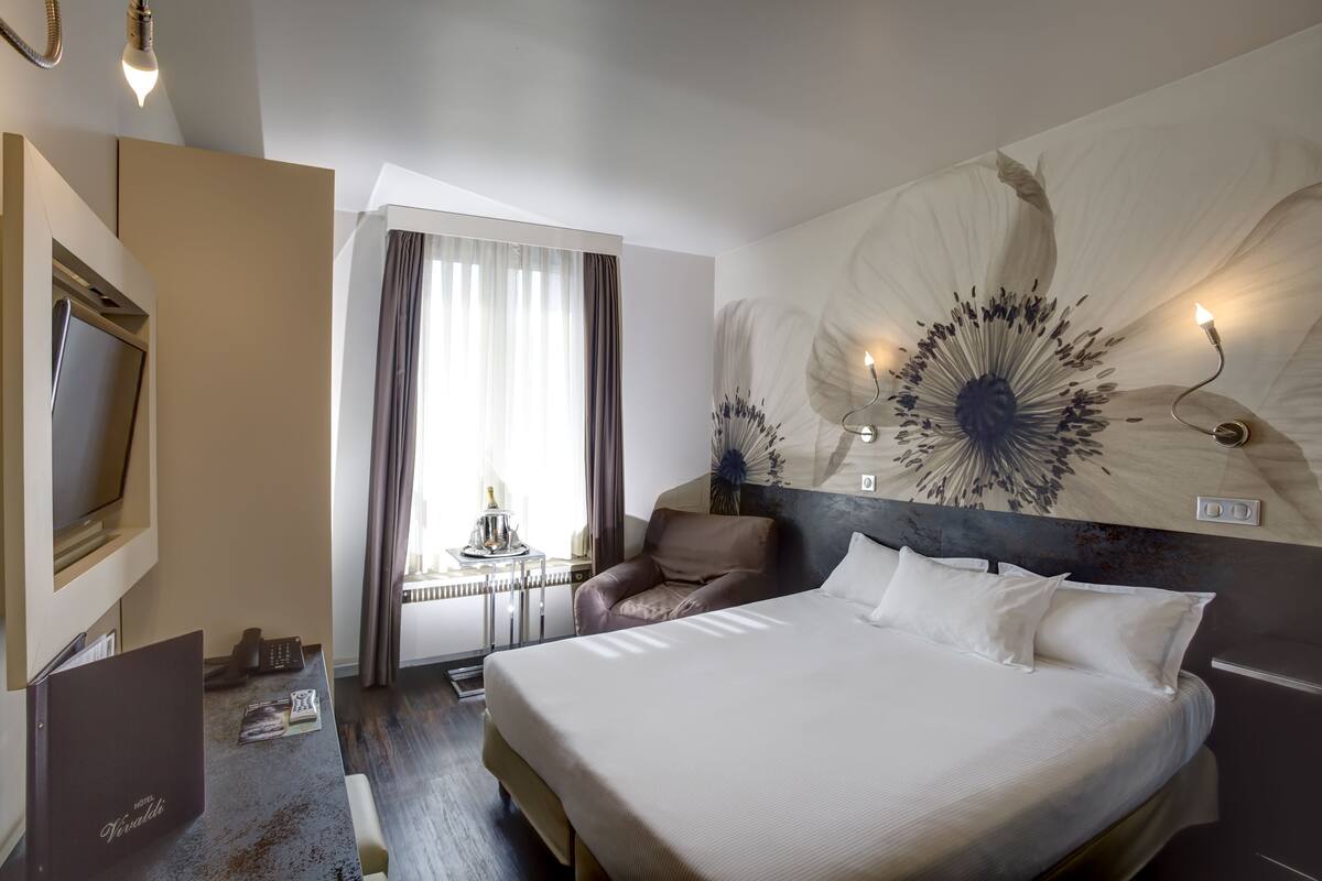 A hotel room with a large bed, a desk with a chair, a television, and a large floral artwork on the wall.