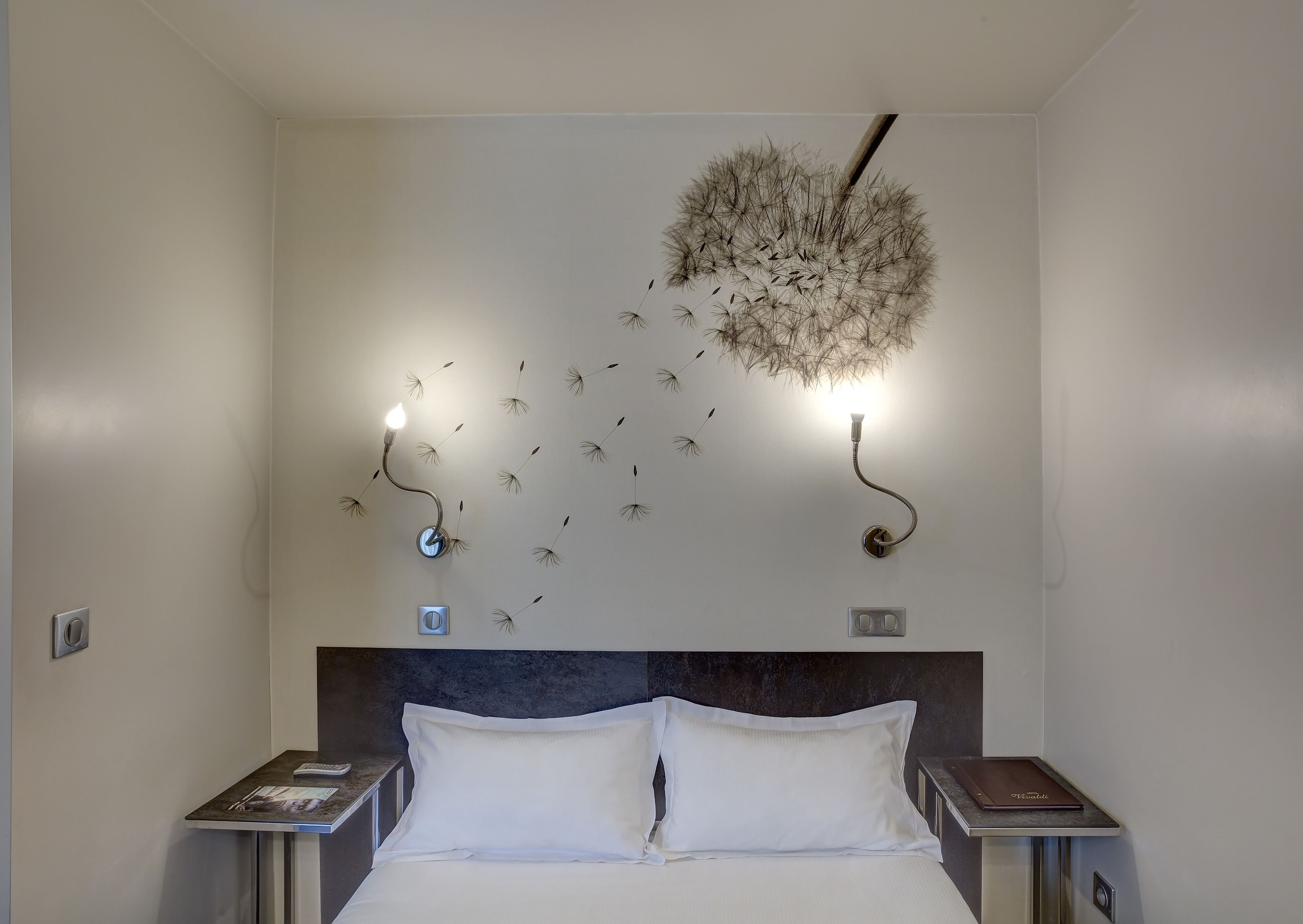 A modern bedroom with a large bed, two bedside tables, and a unique wall art installation featuring a light fixture and dandelion seeds.