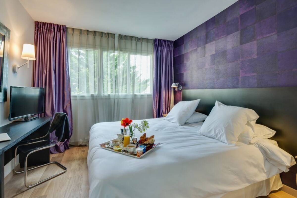 Quarto de hotel com cama, escrivaninha e computador, e bandeja de café da manhã sobre a cama.