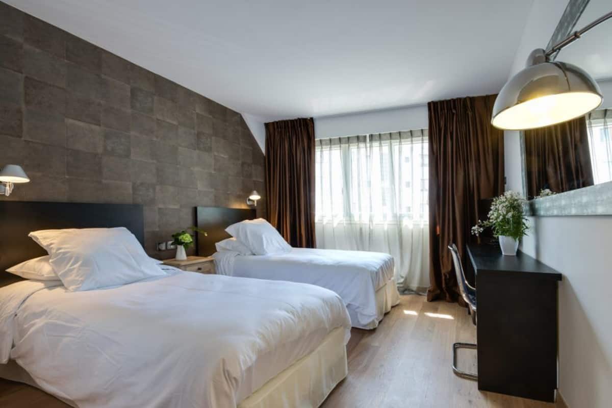 Quarto de hotel com duas camas, uma escrivaninha, uma cadeira, um abajur e uma janela com cortinas.