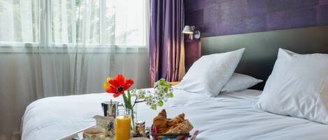 Una cama bien tendida con sábanas blancas, una bandeja de desayuno que incluye croissants, jugo y café, y un jarrón con flores.