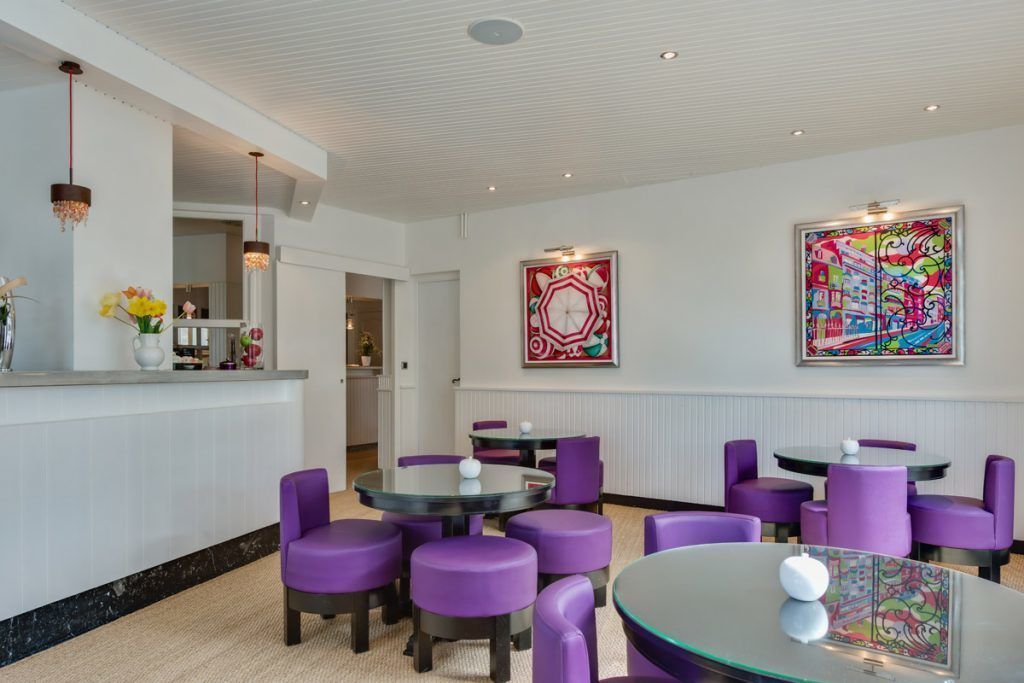 A modern hotel lobby with round tables and purple chairs, a reception desk, and two large abstract paintings on the walls.