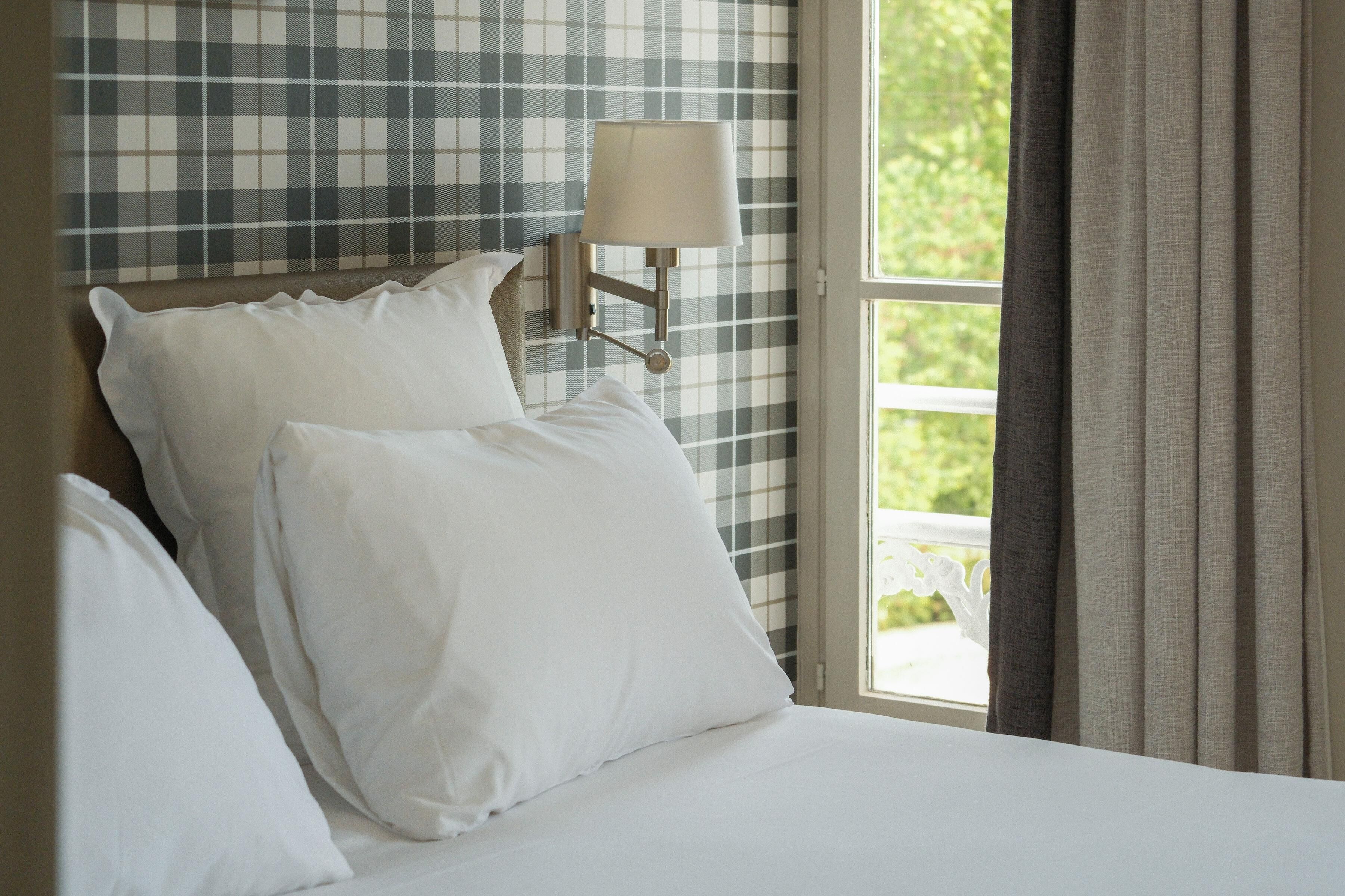 A bed with white linens, a wall-mounted lamp, and a window with curtains.