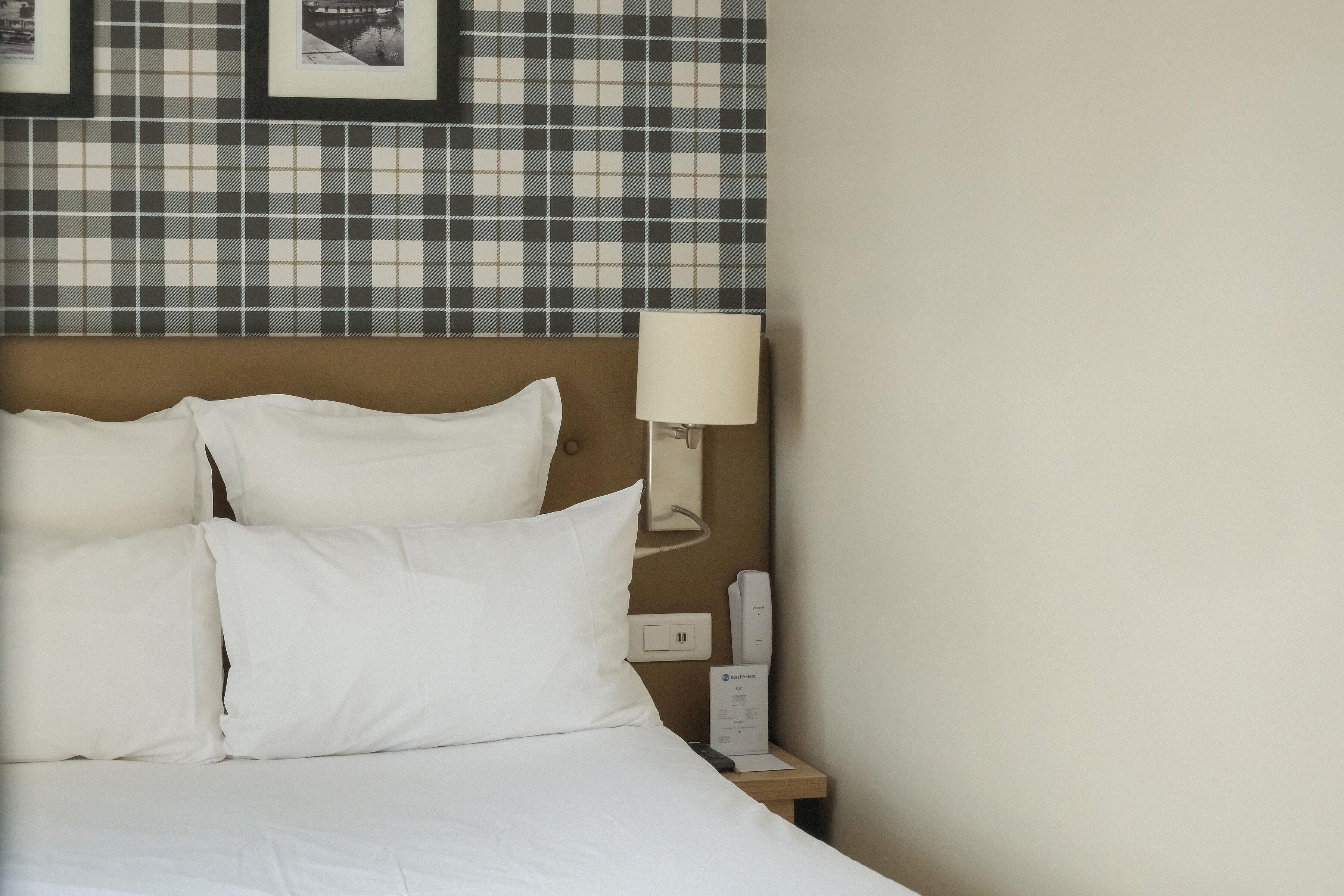 Una habitación de hotel con cama, almohadas, una mesita de noche, lámpara y una pared con diseño a cuadros.