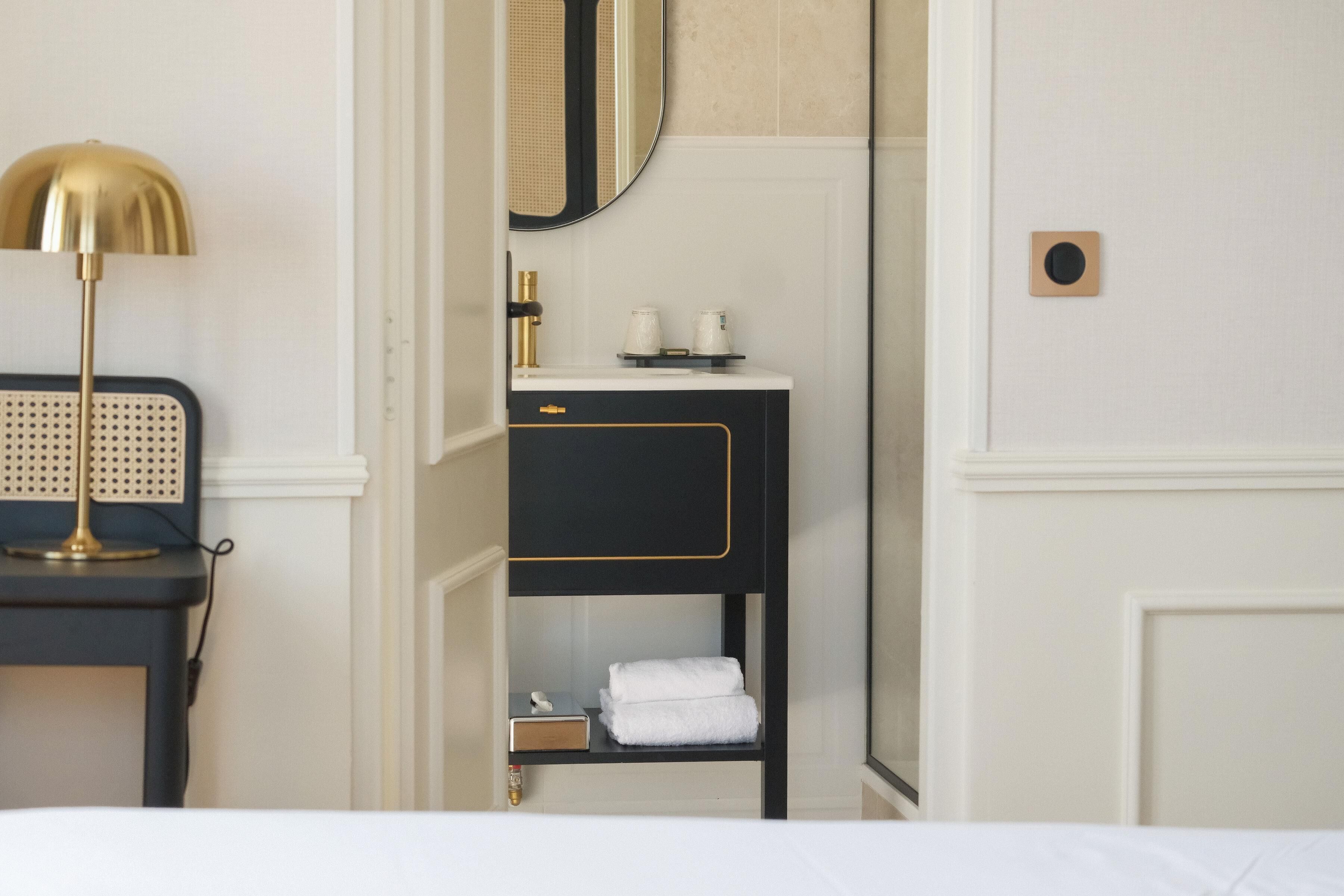 A hotel room with a bed, a bedside table with a lamp, a black and gold vanity with a mirror, and a towel rack.