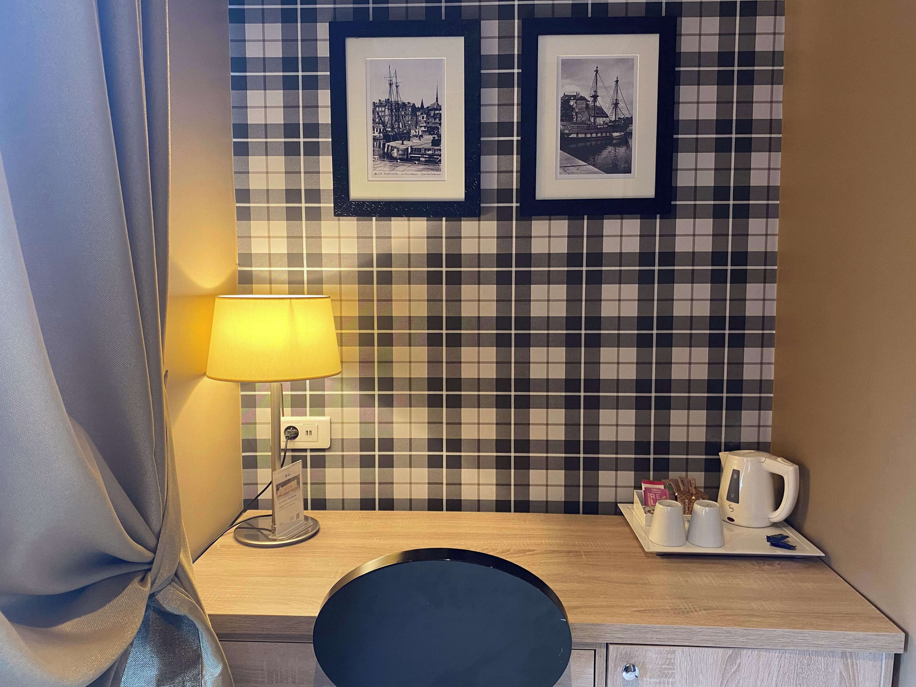 A hotel room with a checkered wall, a desk with a lamp, a coffee maker, and two framed pictures.