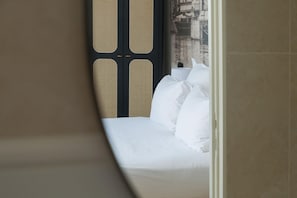 A bed with white linens, a window with a view of buildings, and a door with a woven pattern.
