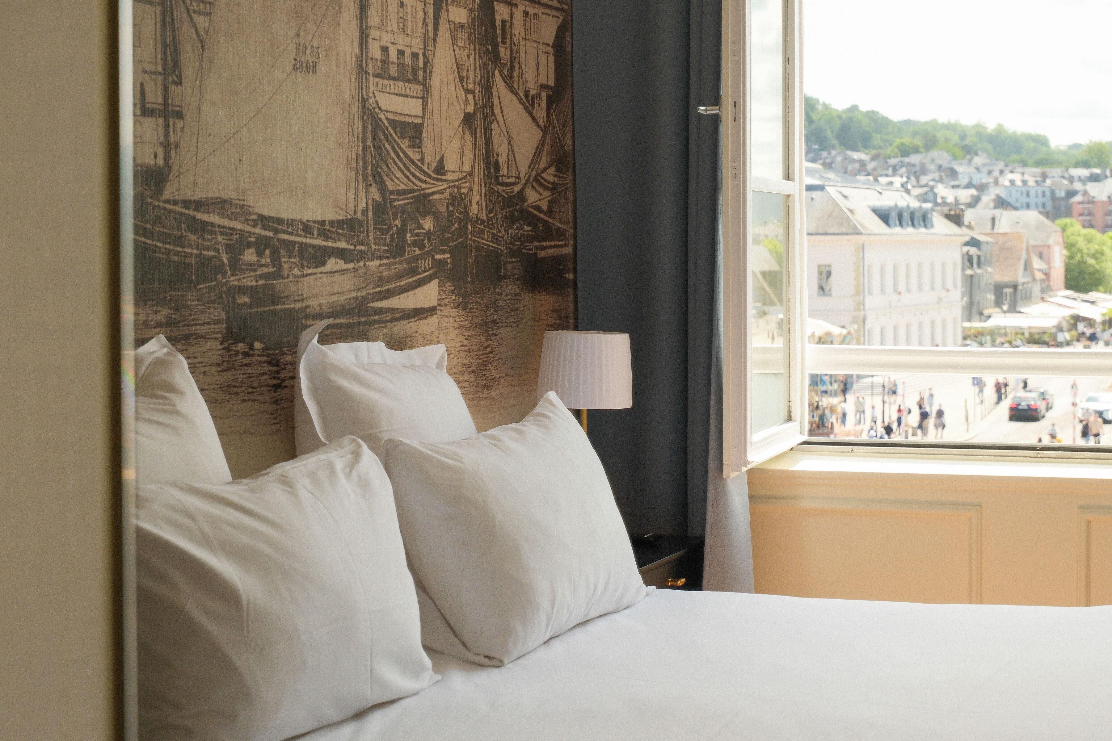 A hotel room with a bed, pillows, a lamp, a large window, and a cityscape view.