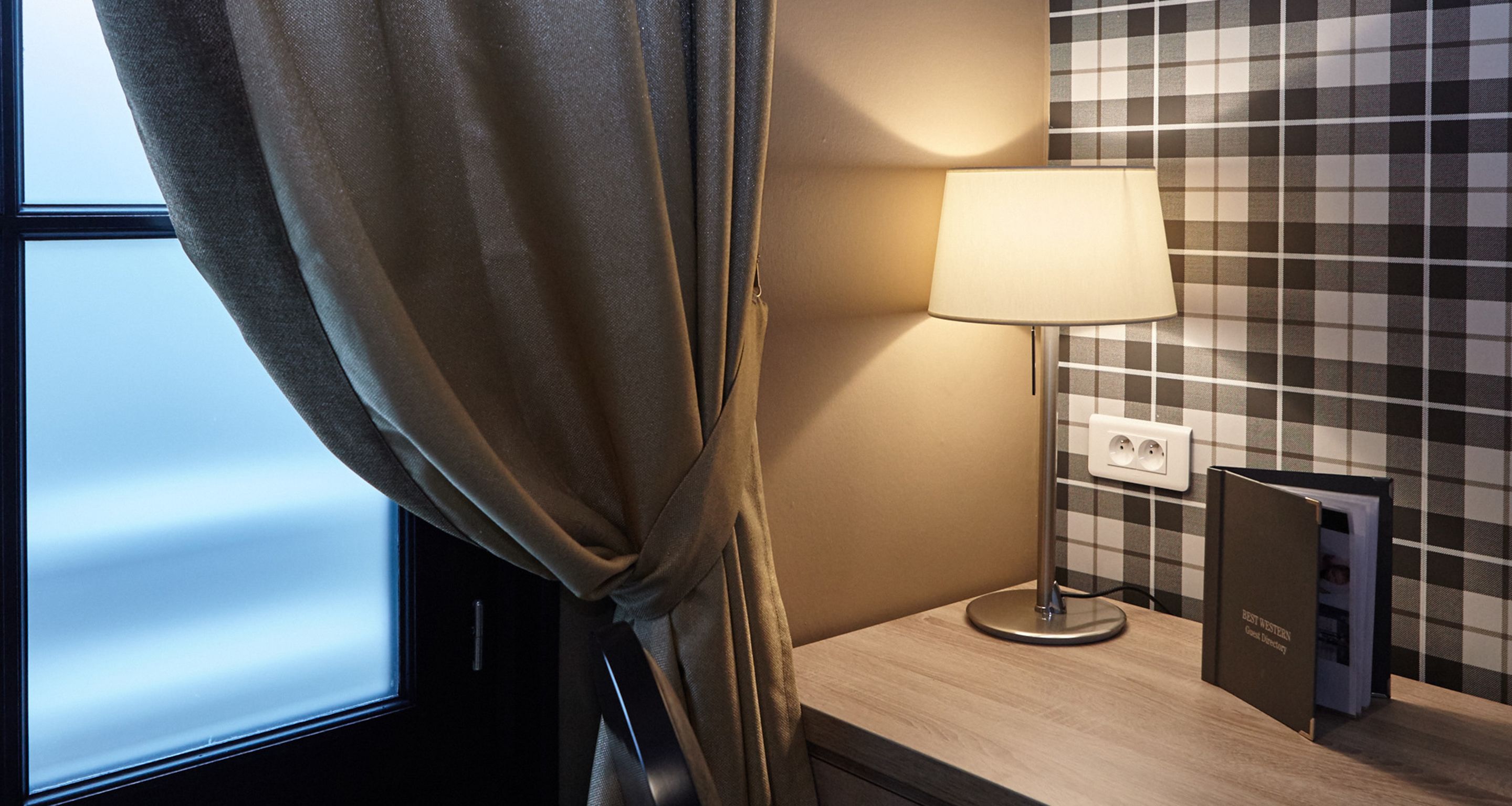 A hotel room with a window, a lamp on a bedside table, and a checkered wall.