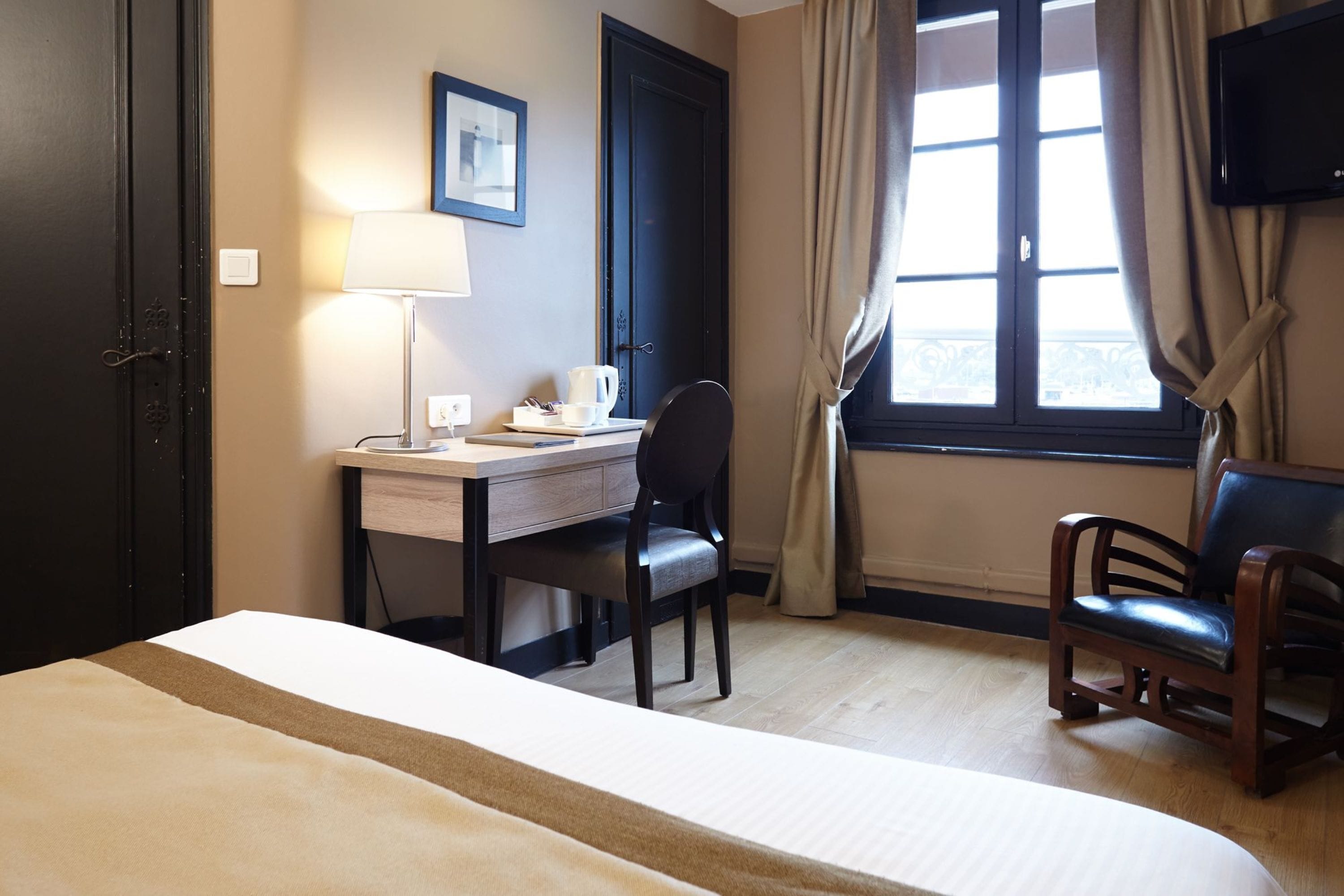 A hotel room with a bed, a desk with a chair, a television, and a window with curtains.