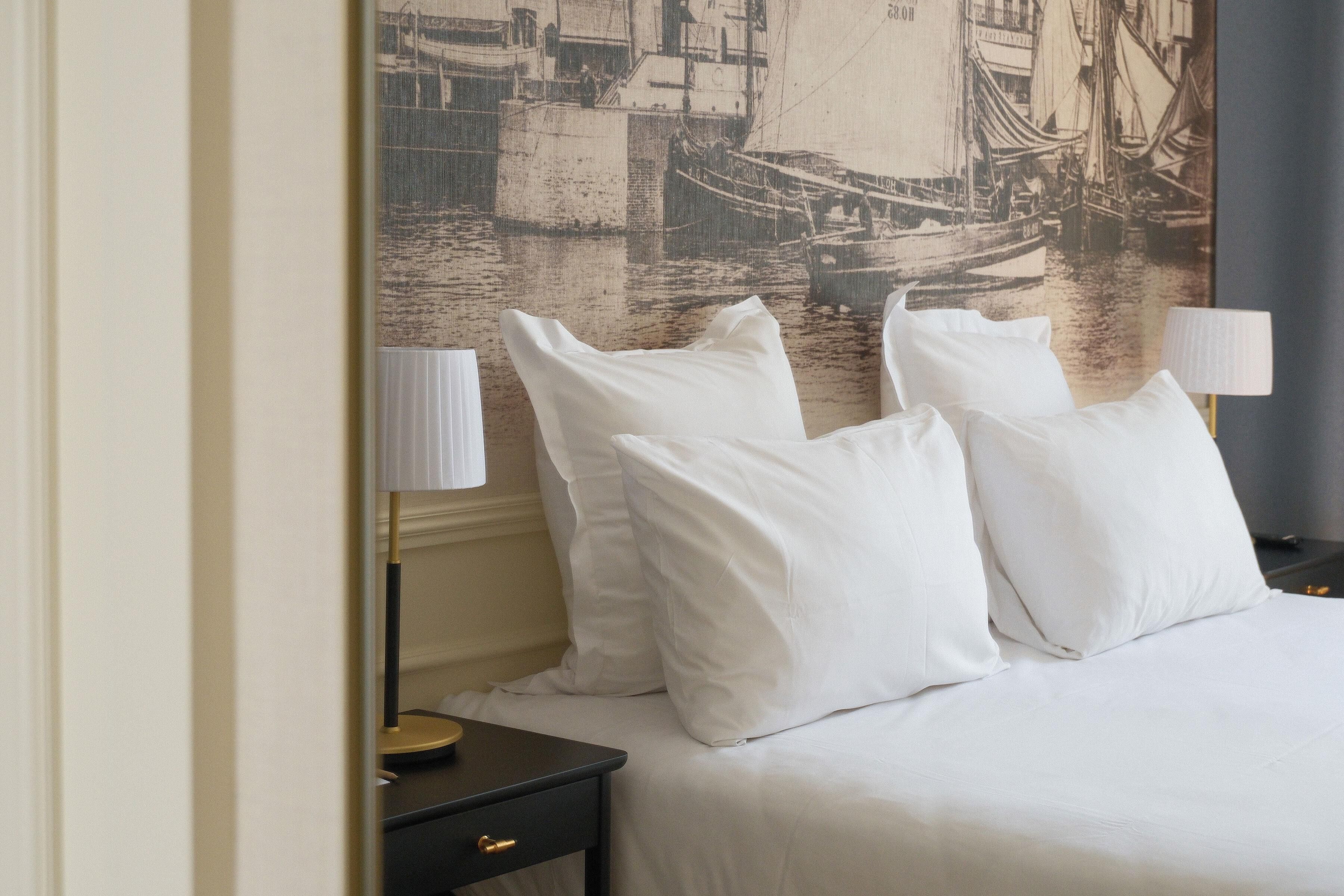 A hotel room with a bed, white pillows, a nightstand, a lamp, and a large maritime-themed wall mural.