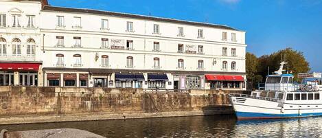 A white building with multiple windows and balconies, a stone wall, and a boat docked at the water's edge.