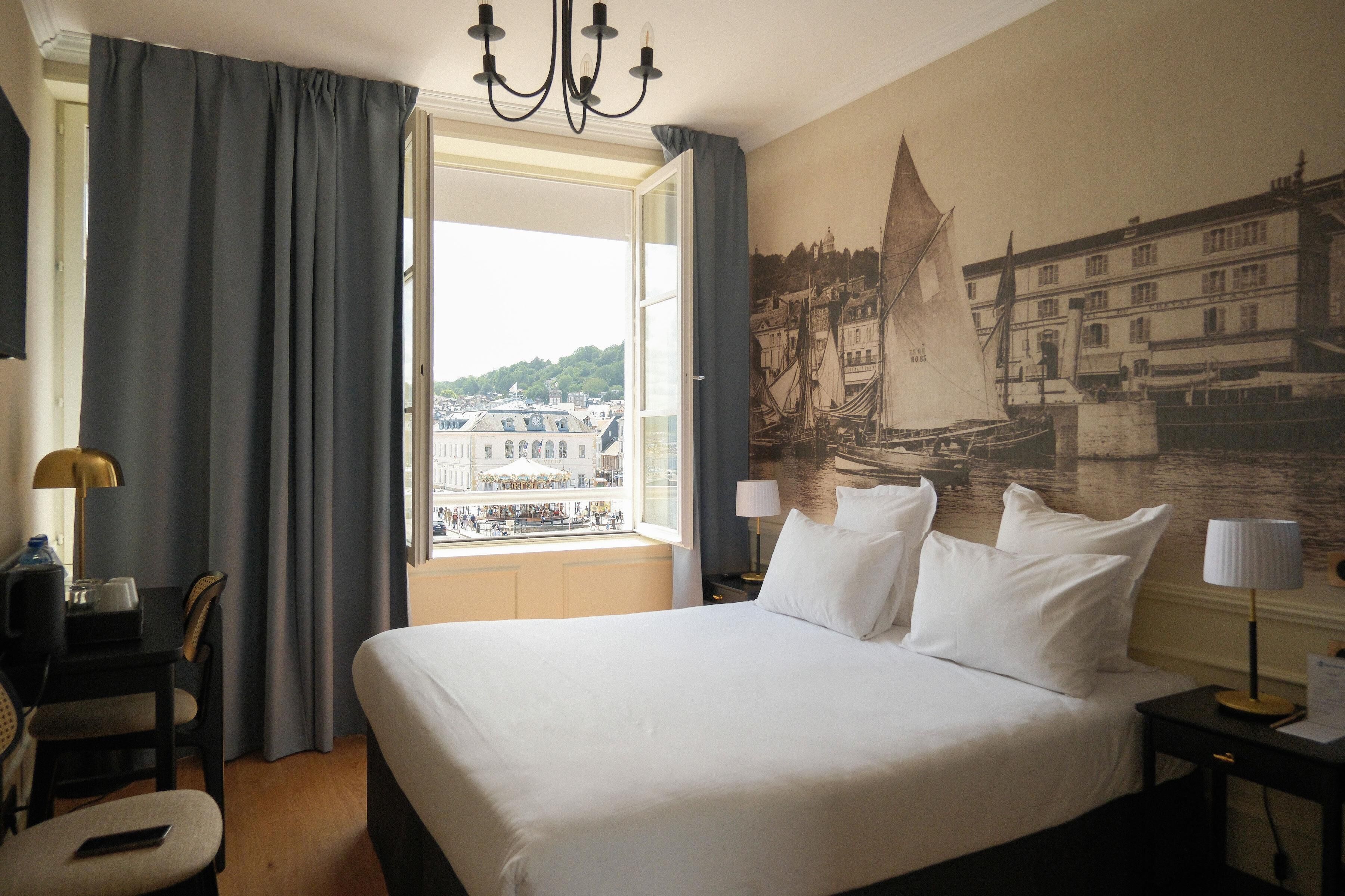 A hotel room with a large bed, a desk, a chair, a lamp, and a view of a cityscape through a window.