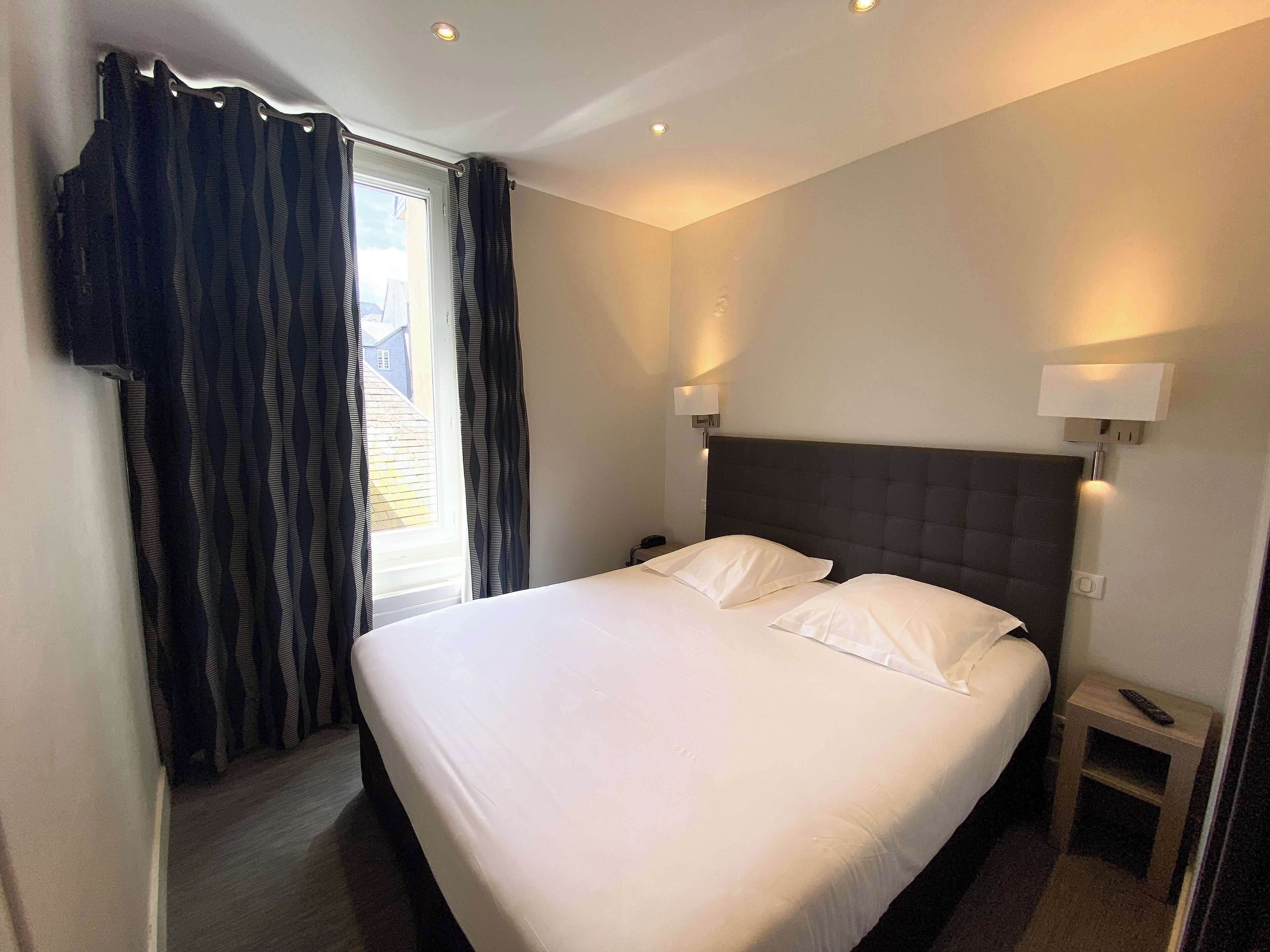 Habitación de hotel con una cama grande, dos mesitas de noche, una ventana con cortinas y una lámpara fijada a la pared.