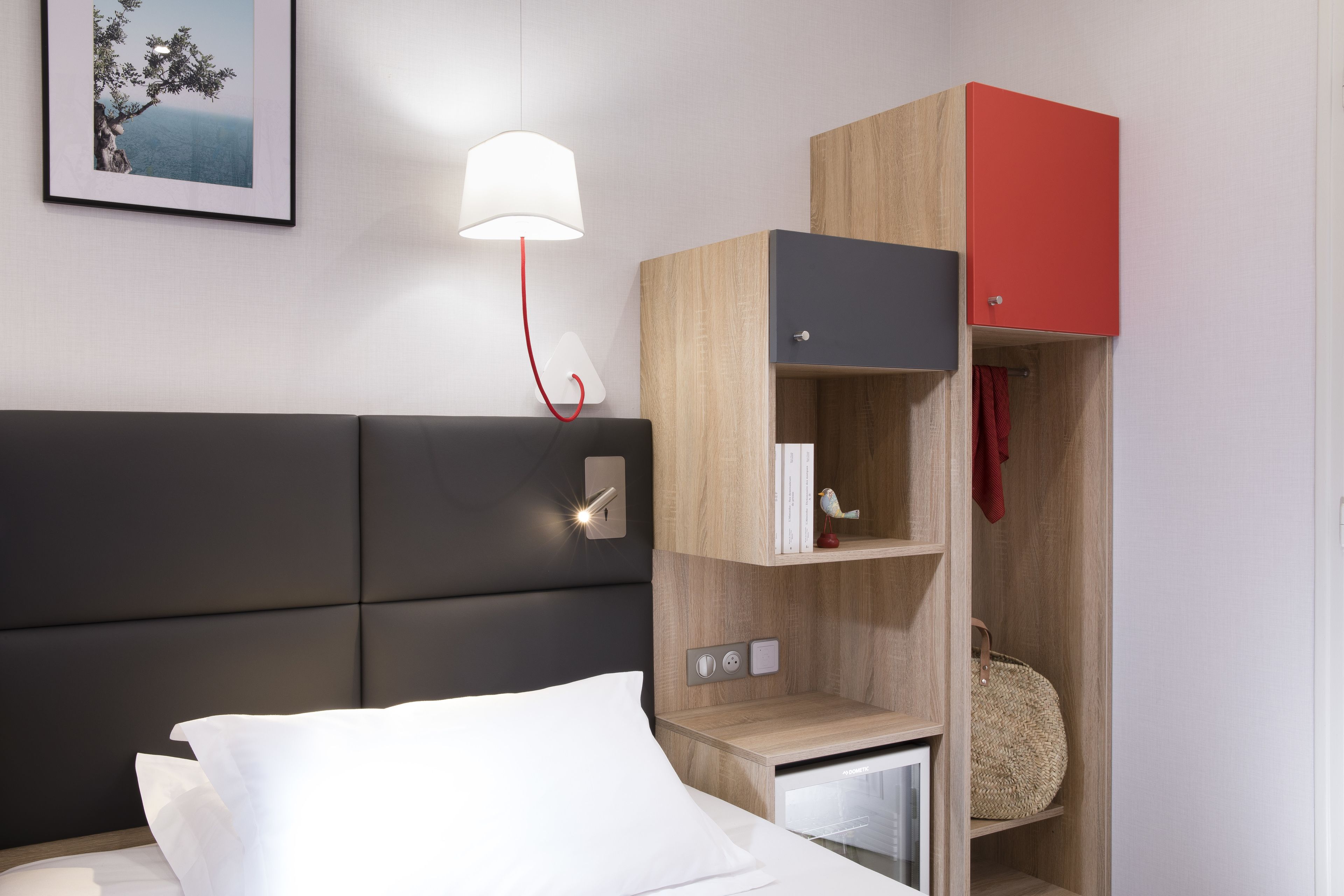 A modern hotel room with a bed, a nightstand, a lamp, a wardrobe, and a framed picture on the wall.