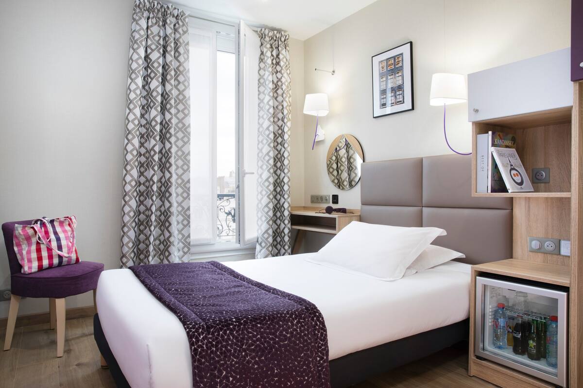 Habitación de hotel con cama, una silla, mesita de noche con lámpara, una pequeña mesa y un mini refrigerador.