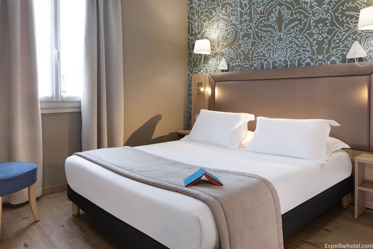Una habitación de hotel con una cama, una mesita de noche, una lámpara y una ventana con cortinas.