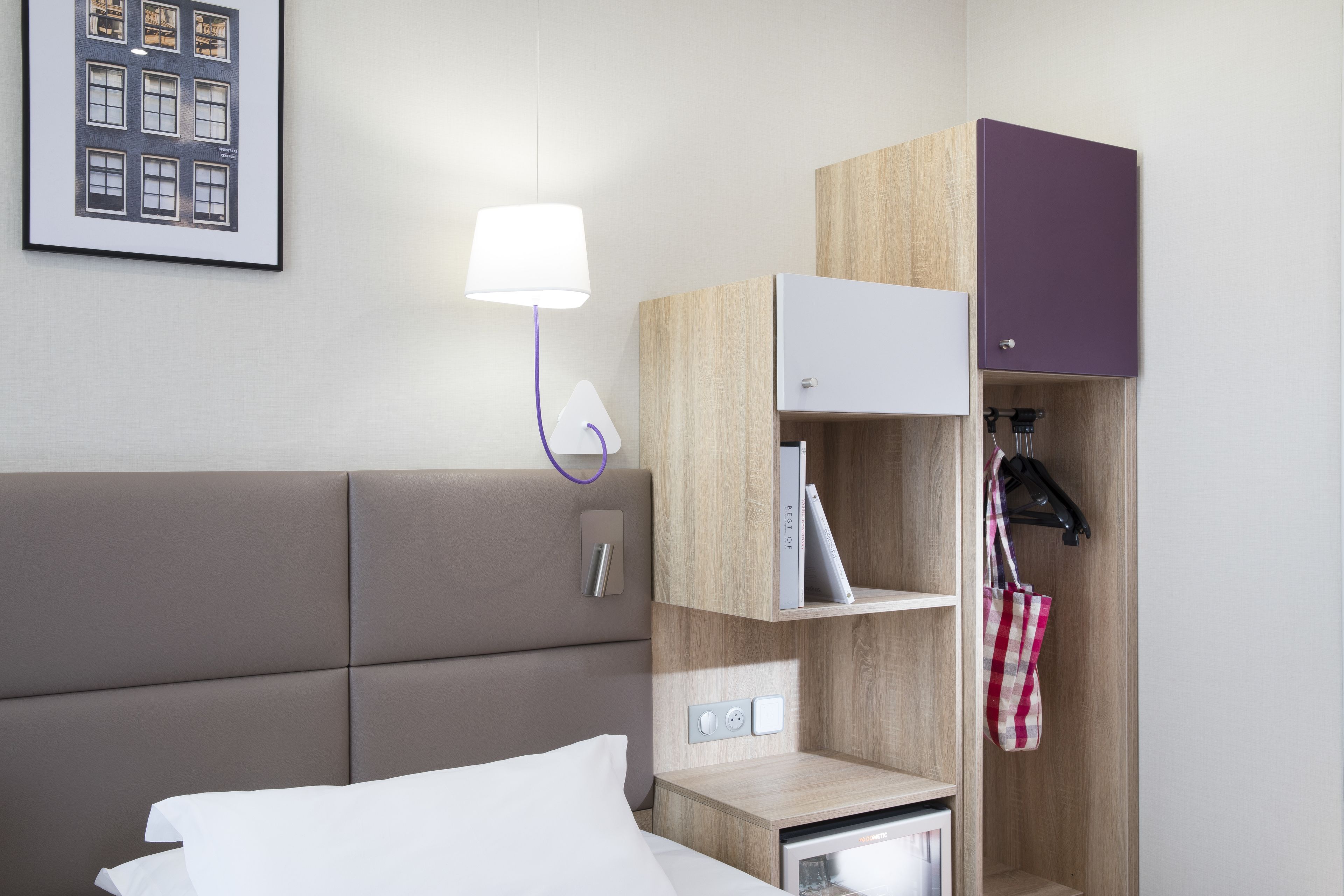 A hotel room with a bed, a bedside table with a lamp, a small cabinet, and a coat rack with a bag.