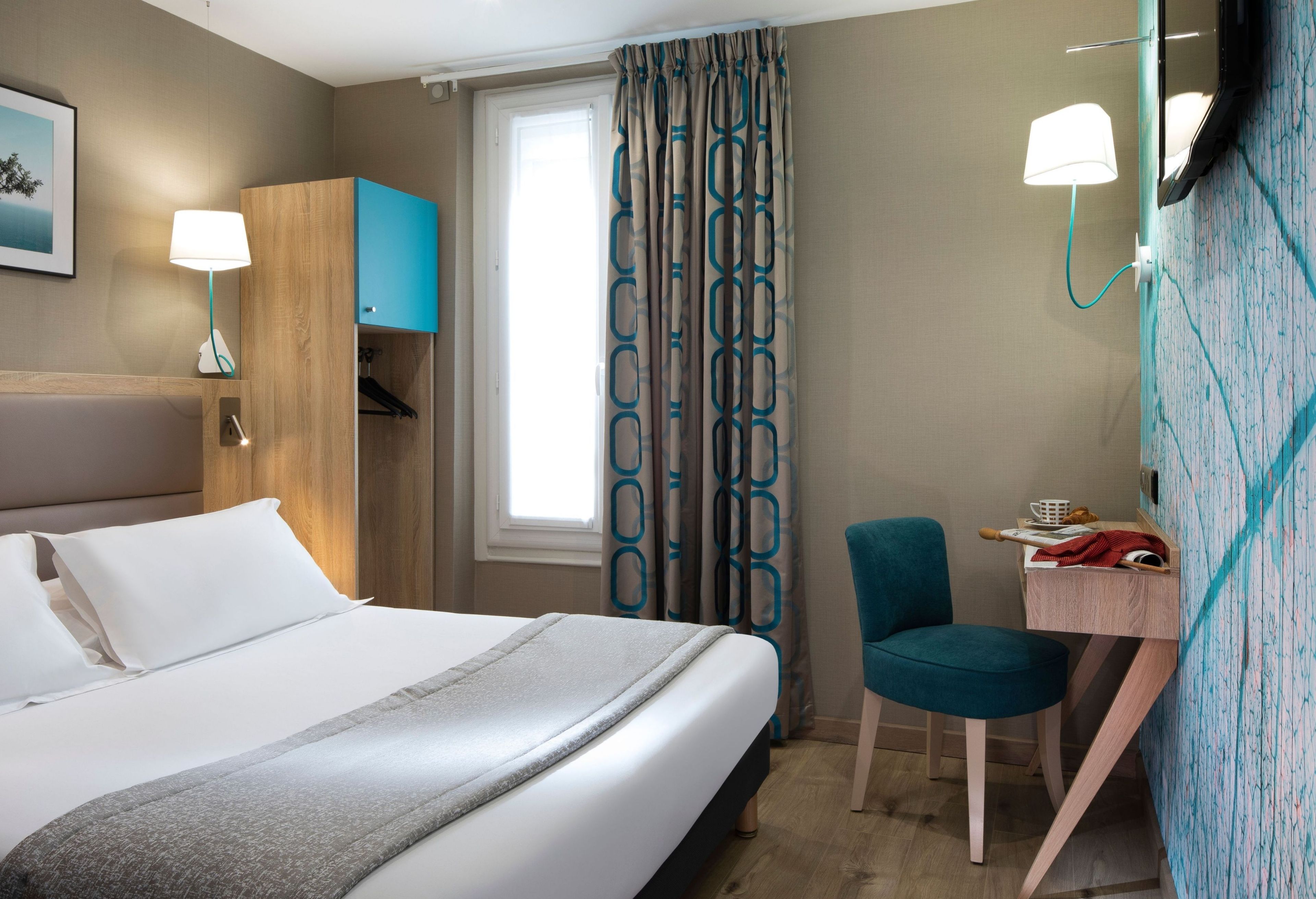 A hotel room with a bed, a desk, a chair, a wardrobe, a window with curtains, and a wall-mounted lamp.