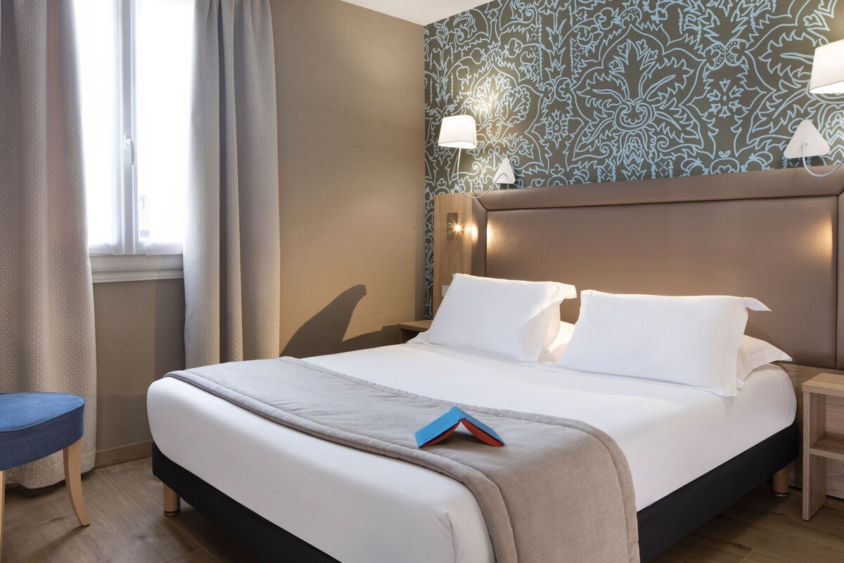 A hotel room with a bed, a nightstand, a lamp, and a window with curtains.