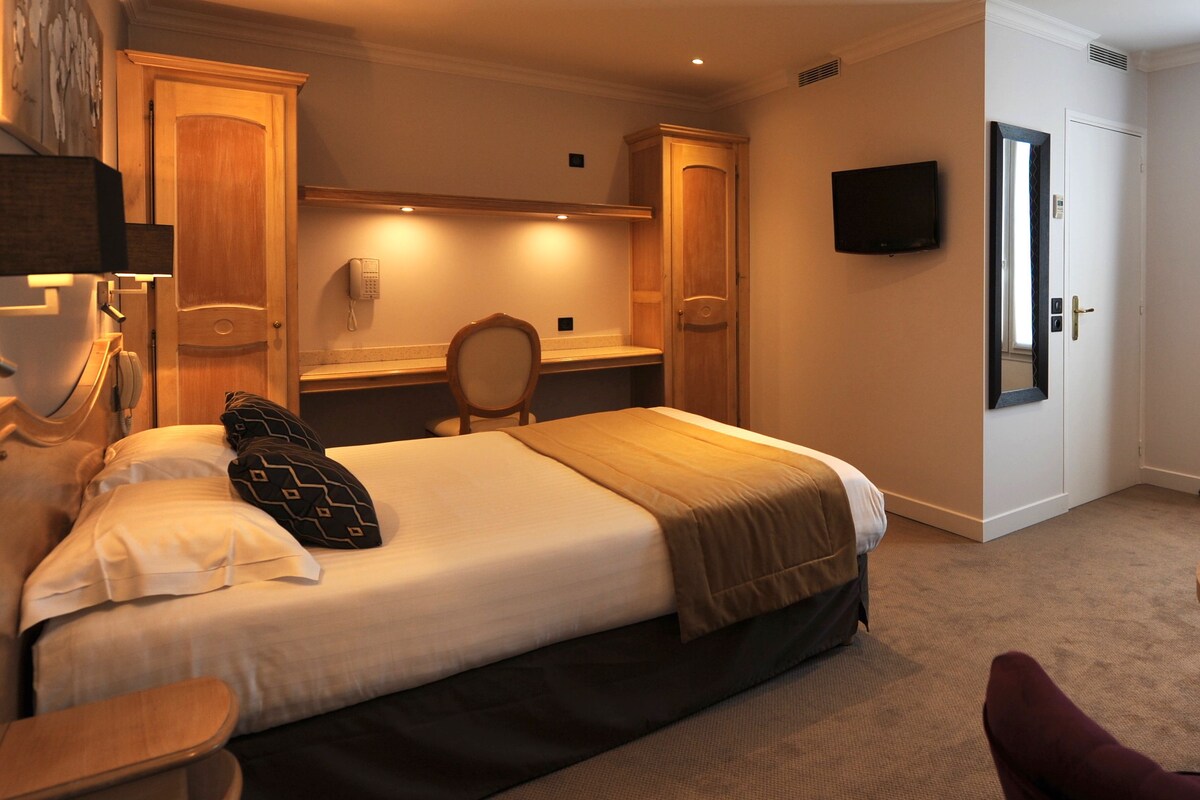 Habitación de hotel con cama, escritorio, silla, mesa redonda, televisor y puerta con cortinas.