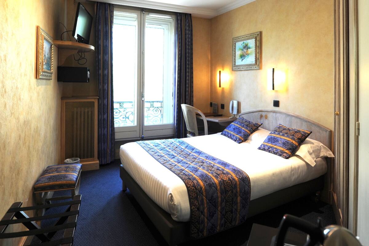 A hotel room with a bed, a desk, a chair, a television, and a balcony with curtains.