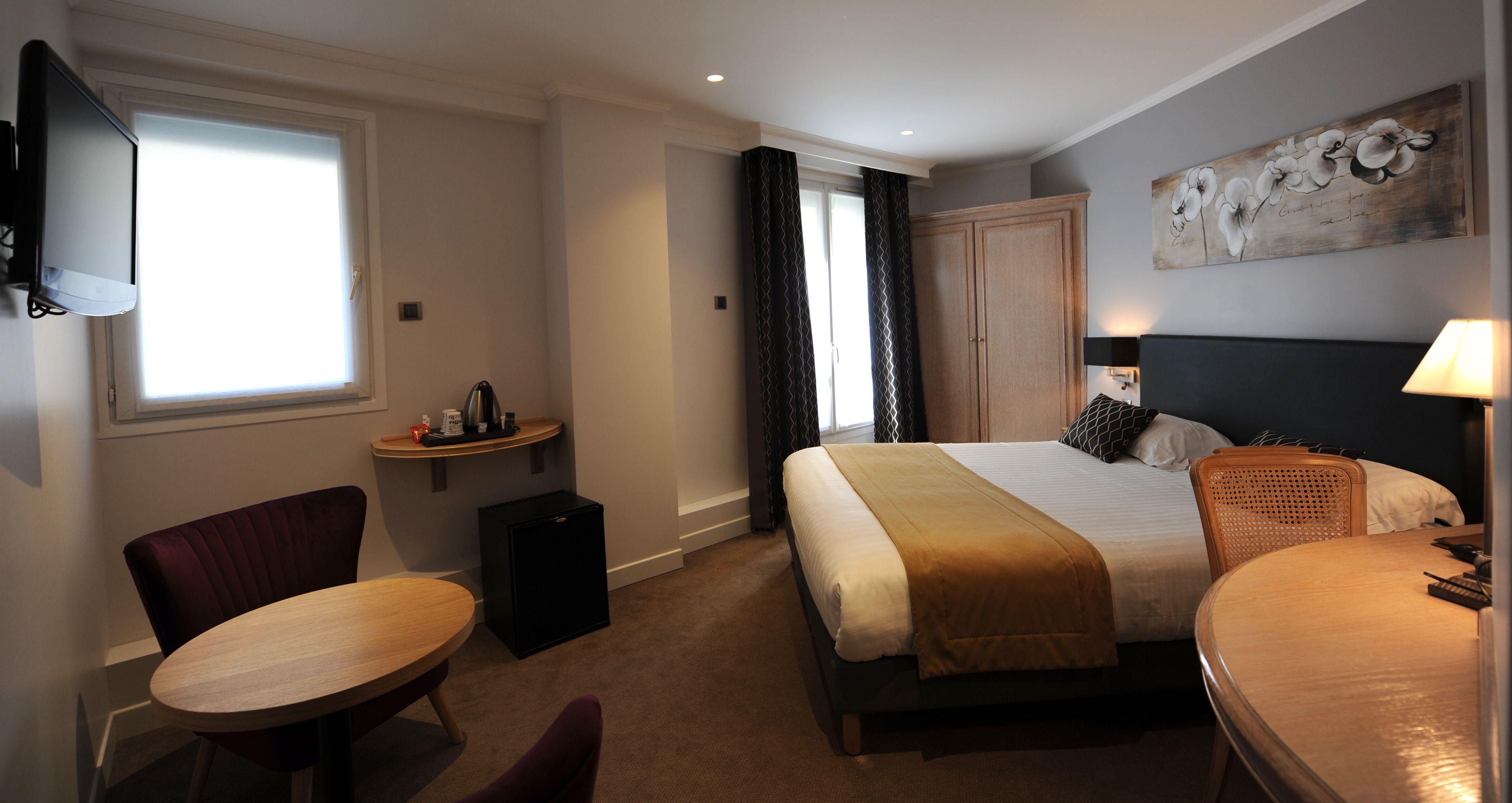 A hotel room with a bed, a desk, a chair, a small table, a TV, a wardrobe, and a painting on the wall.