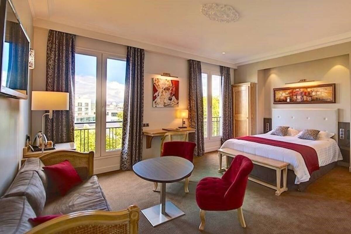 A hotel room with a bed, a sofa, a dining table, and a chair. There is a window with curtains, a painting on the wall, and a lamp.