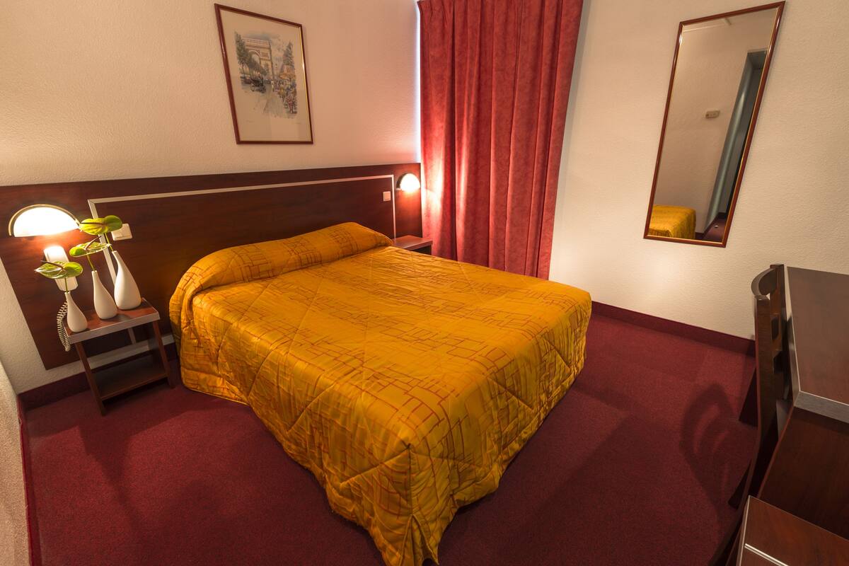 A hotel room with a bed, a nightstand, a lamp, a mirror, and a red curtain.