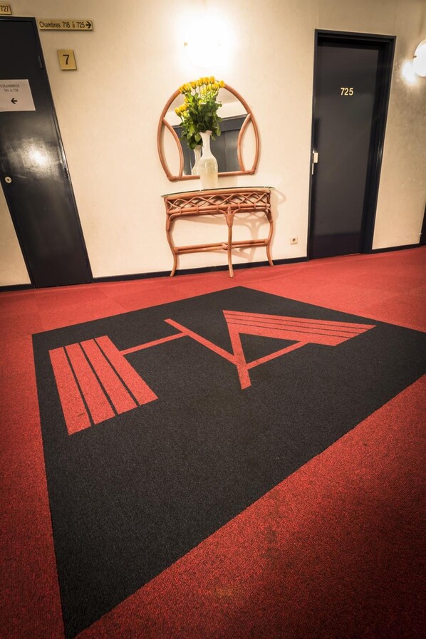 A hotel corridor with a red carpet, a mirror, a vase with yellow flowers, and a door labeled 725.