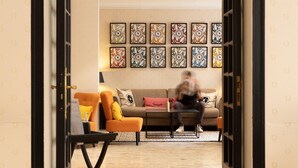A modern living room with a sofa, armchairs, and a coffee table. There are framed artworks on the wall and a door leading to another room.