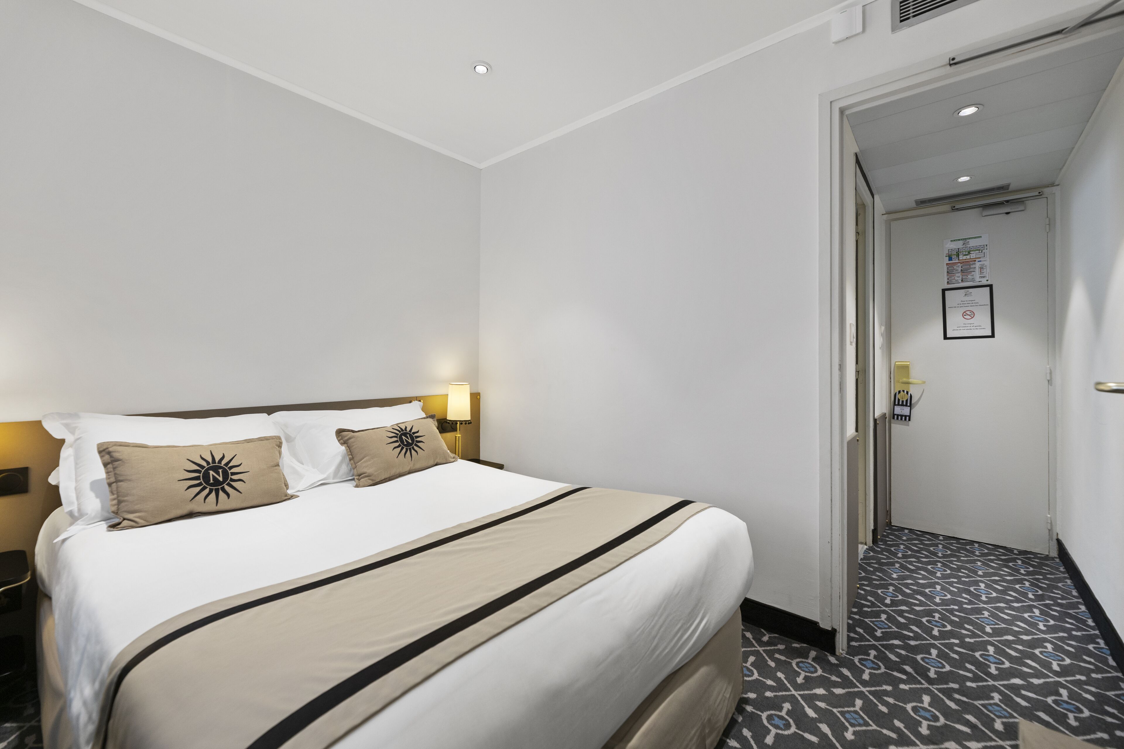 A hotel room with a neatly made bed, a nightstand, a lamp, and a door leading to a hallway.