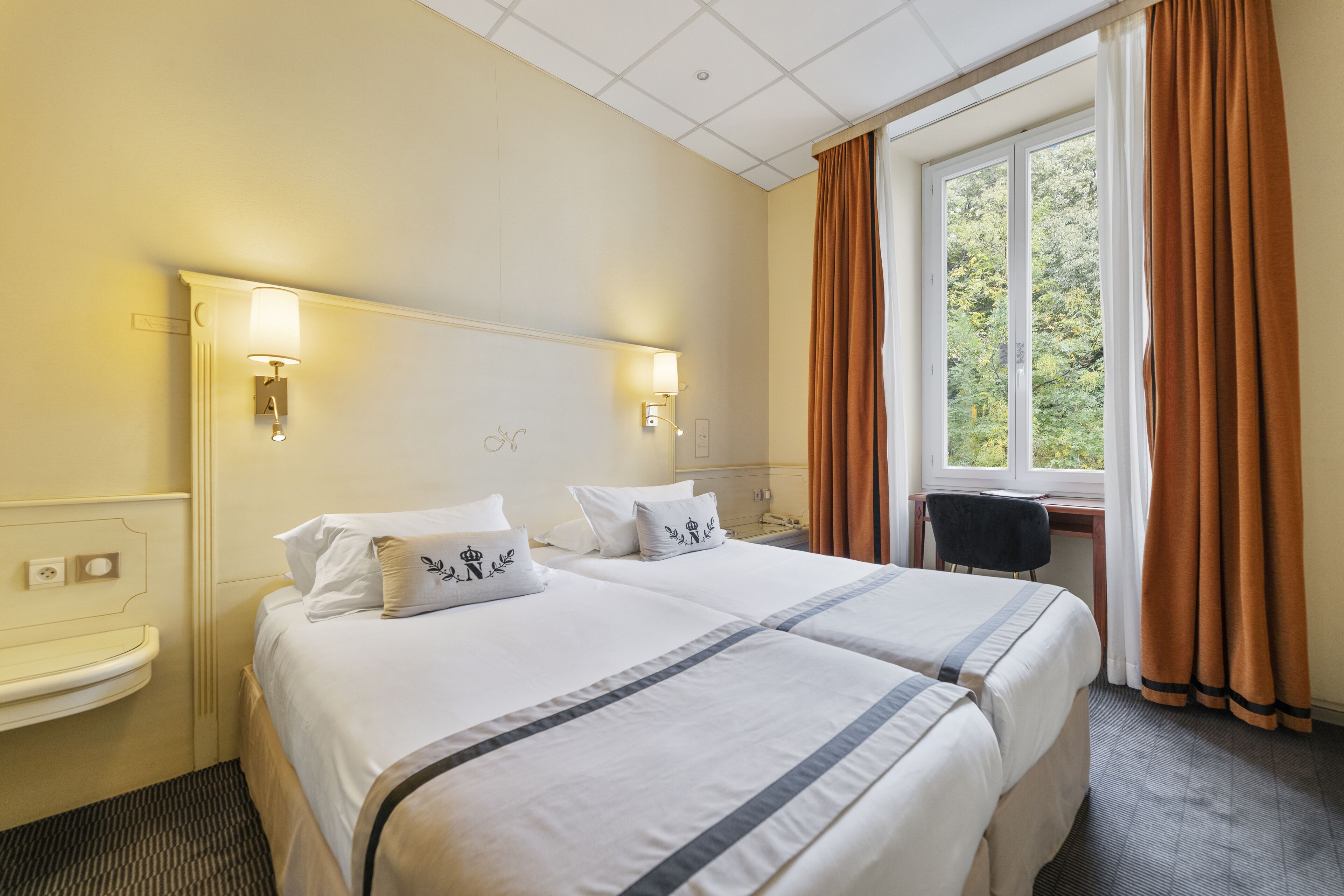 A hotel room with two beds, a bedside table, a desk, and a window with curtains.