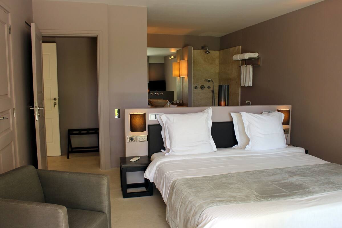 A hotel room with a bed, a chair, a nightstand, a mirror, and a bathroom visible through an open door.