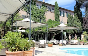 A poolside area with outdoor seating, umbrellas, and a building in the background. - Le Mas de Chastelas (Gassin)