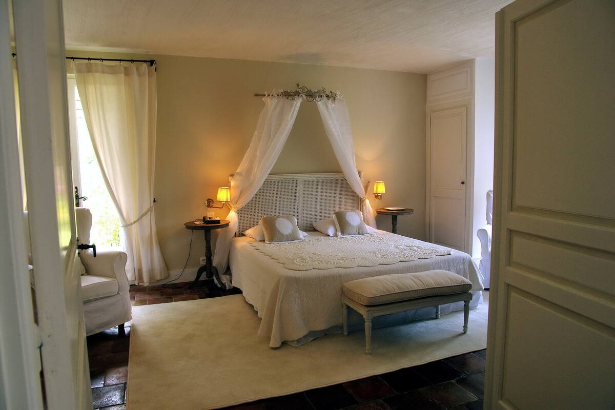 A bedroom with a canopy bed, bedside lamps, a chair, and a bench.