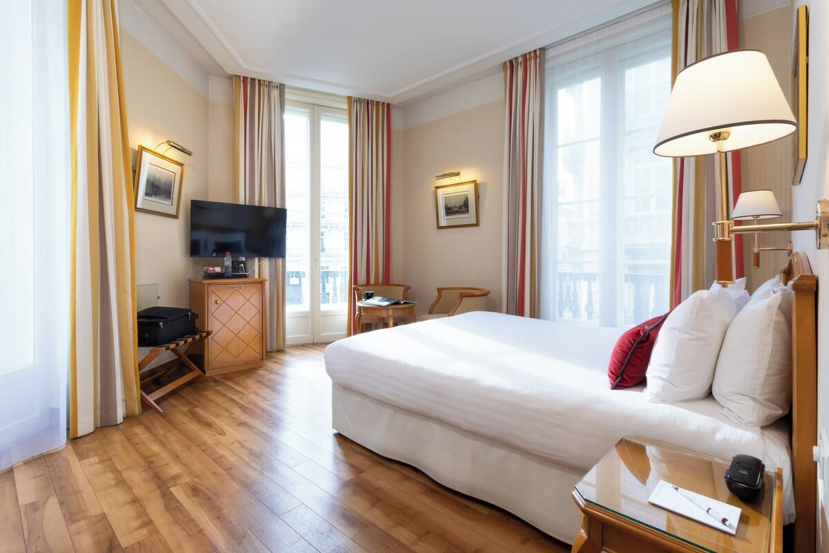 A hotel room with a large bed, a TV, a desk with a chair, and a window with curtains.
