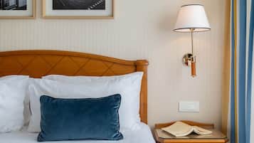 Una cama bien hecha con almohadas blancas y azules, un cabecero de madera, una mesita de noche con un libro y una lámpara fijada a la pared.